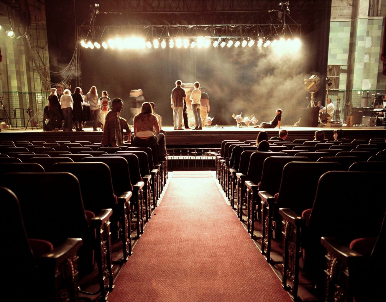 View down central aisle of a theatre towards stage where actors and creative performers are rehearsing.